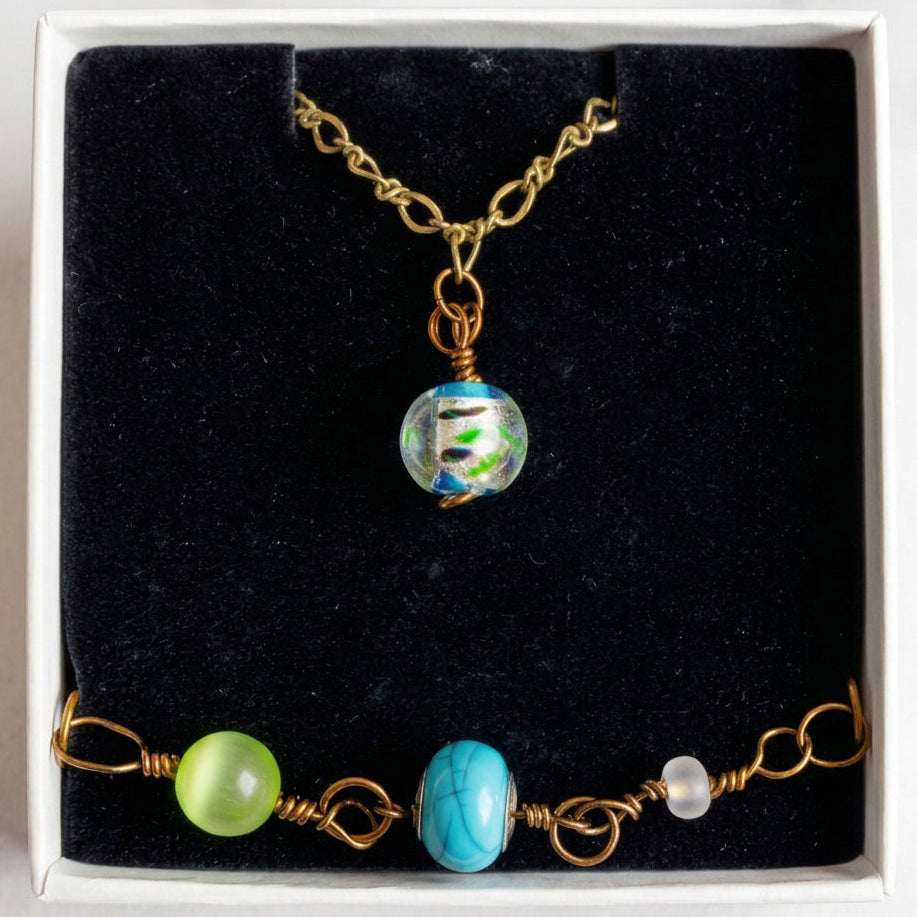 A handmade jewelry set displayed inside a white box with a black velvet lining, featuring a gold chain necklace with a clear glass bead and a matching gold wire-wrapped bracelet with green, blue, and white beads.