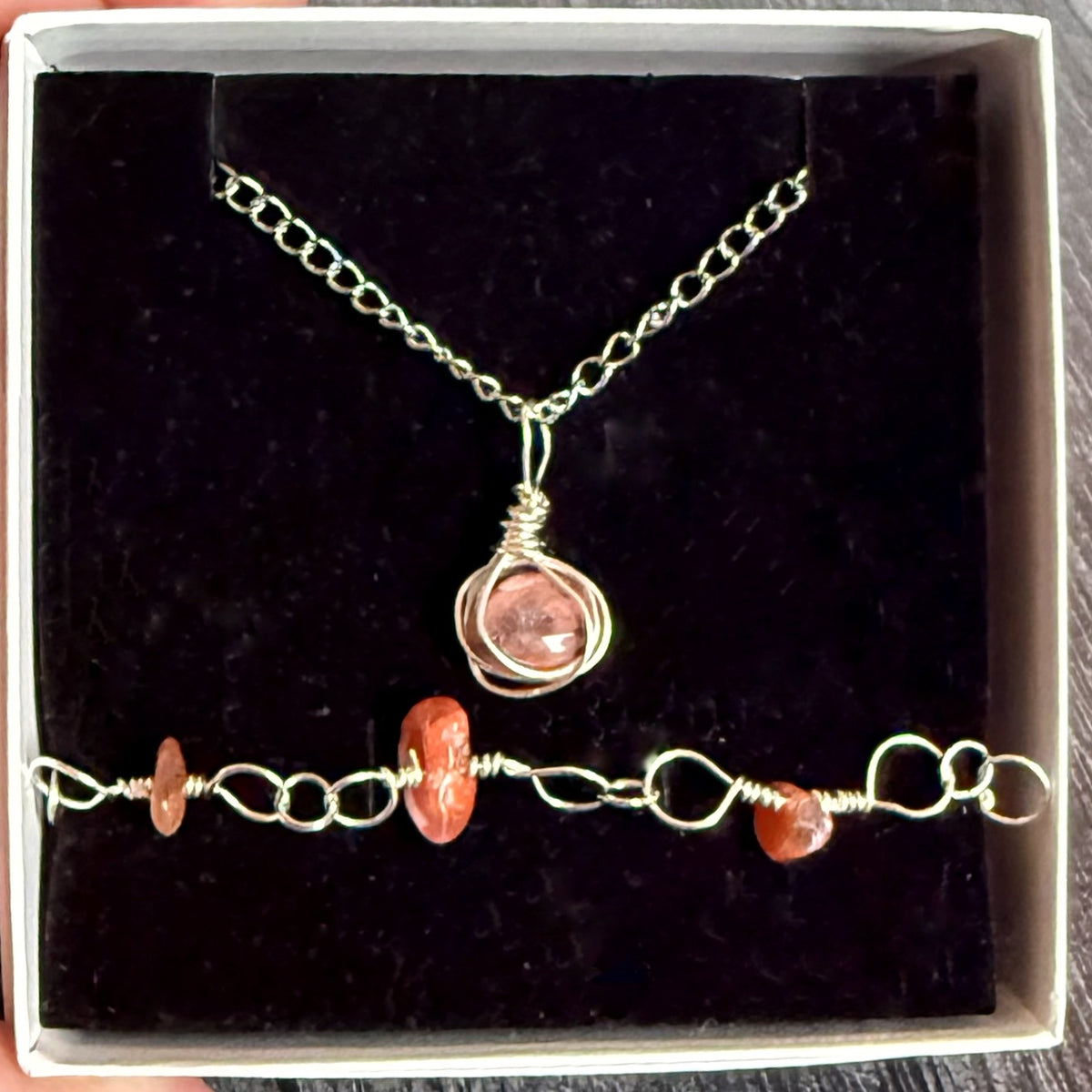 A handmade jewelry set featuring a silver chain necklace with a pink stone wrapped in wire and a matching silver beaded bracelet, displayed on a black velvet insert inside a white gift box.