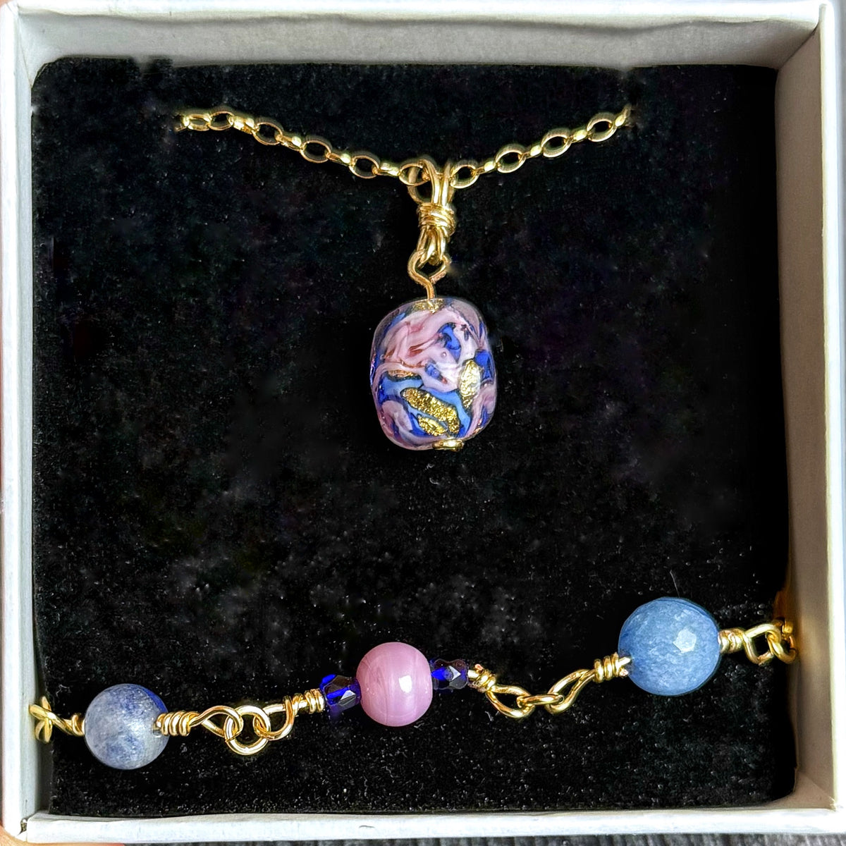 A jewelry set displayed in a white gift box with a black velvet insert, featuring a gold-tone chain necklace with a colorful marbled pendant and a matching beaded bracelet with blue, pink, and gold beads.