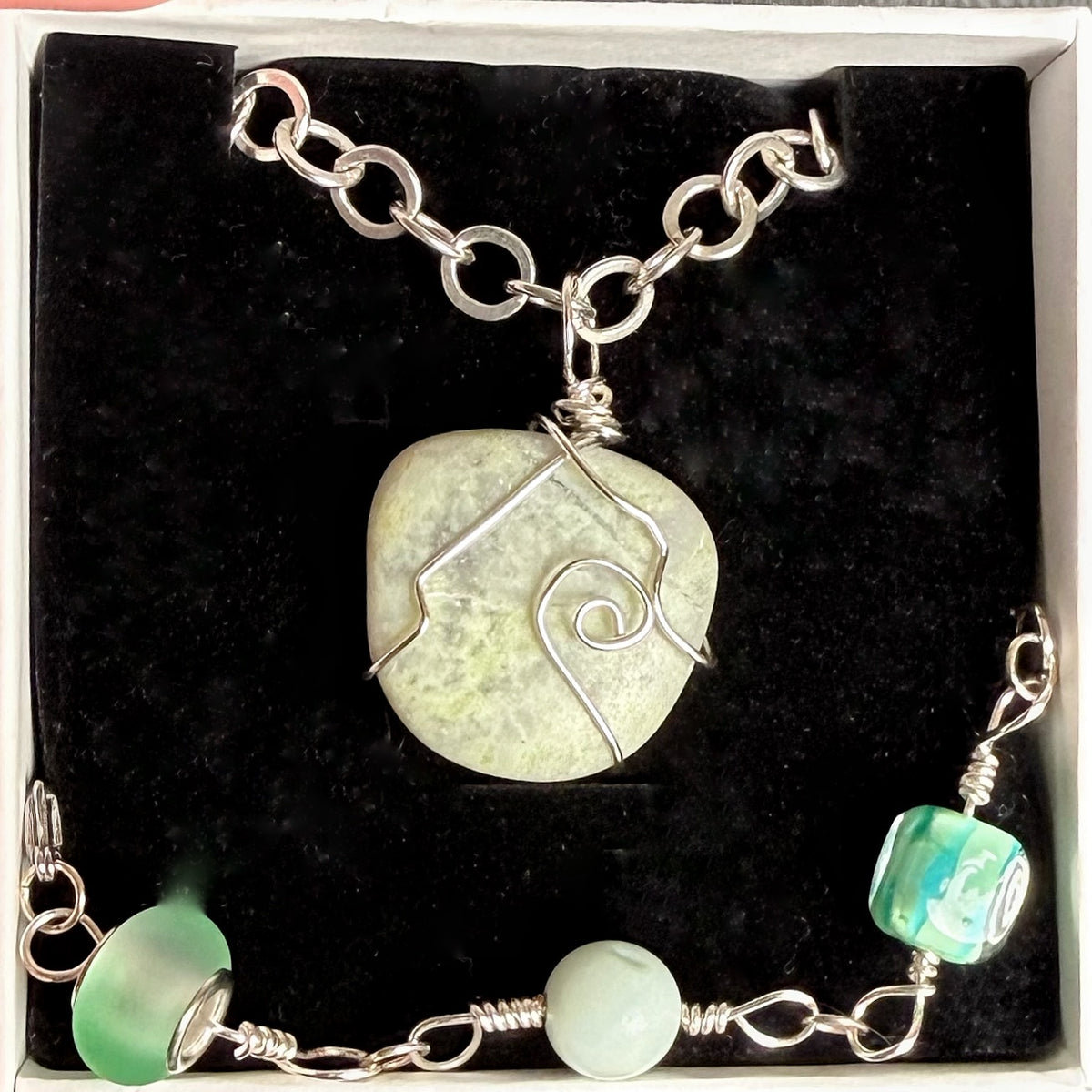 A handmade jewelry set displayed in a black velvet box, featuring a silver chain necklace with a light green stone pendant and a matching beaded bracelet.