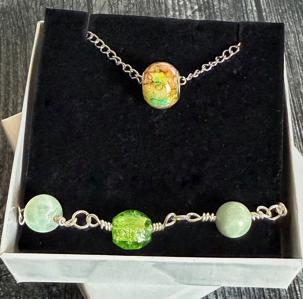 A jewelry set displayed inside an open white box with a black velvet insert, featuring a silver-tone chain necklace with a round multicolor bead and a matching beaded bracelet with pale green and bright green beads.