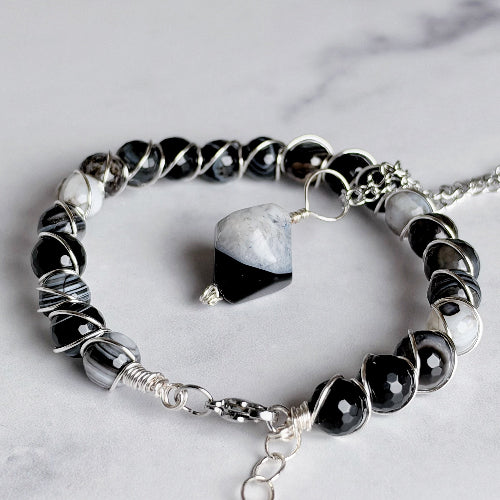 A beaded bracelet with faceted black and white beads, wrapped in silver wire, a silver clasp, and a central polished black-and-white stone, photographed on a light marble surface.