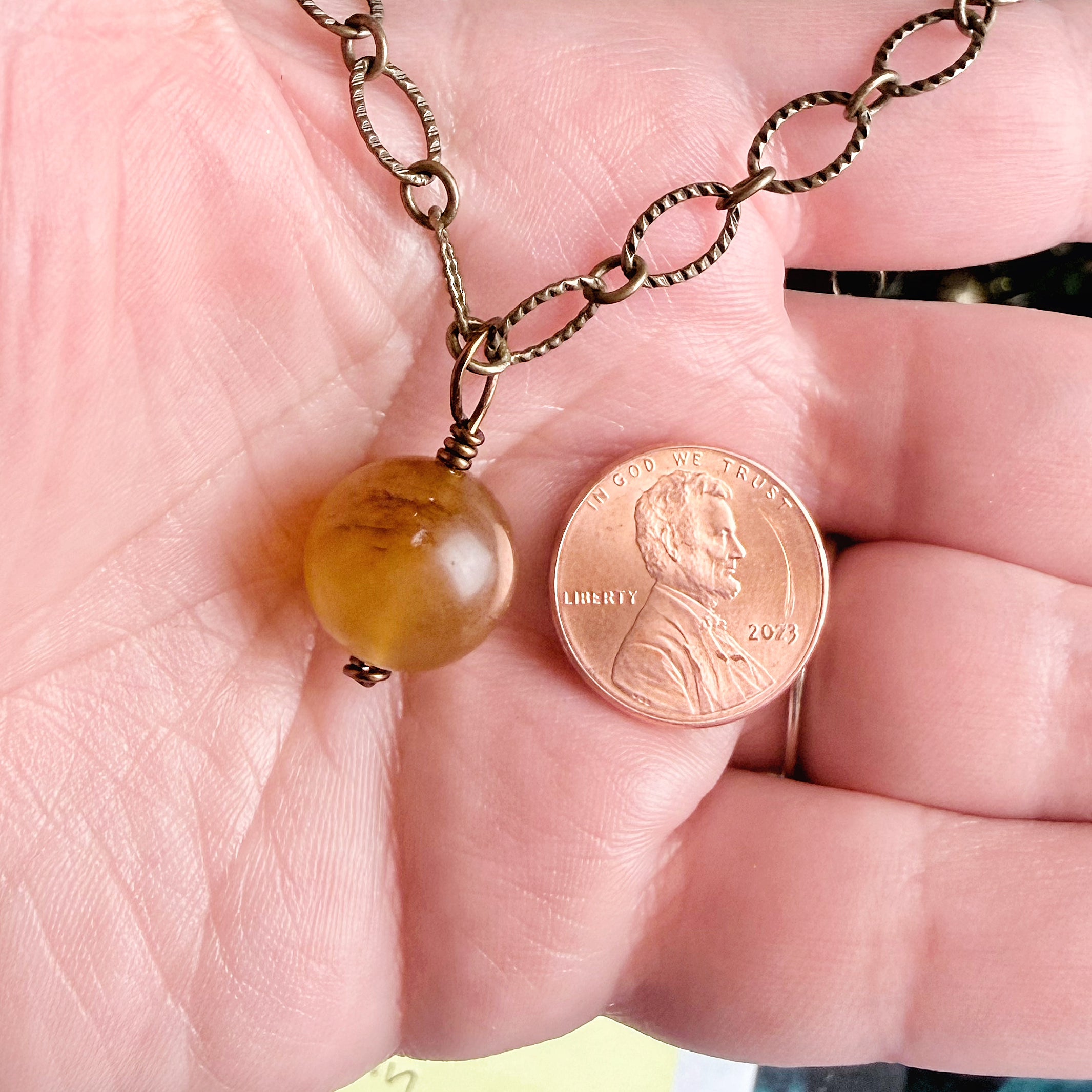 A close-up of a hand holding a bronze chain necklace with a round amber-yellow bead pendant next to a U.S. penny for scale.