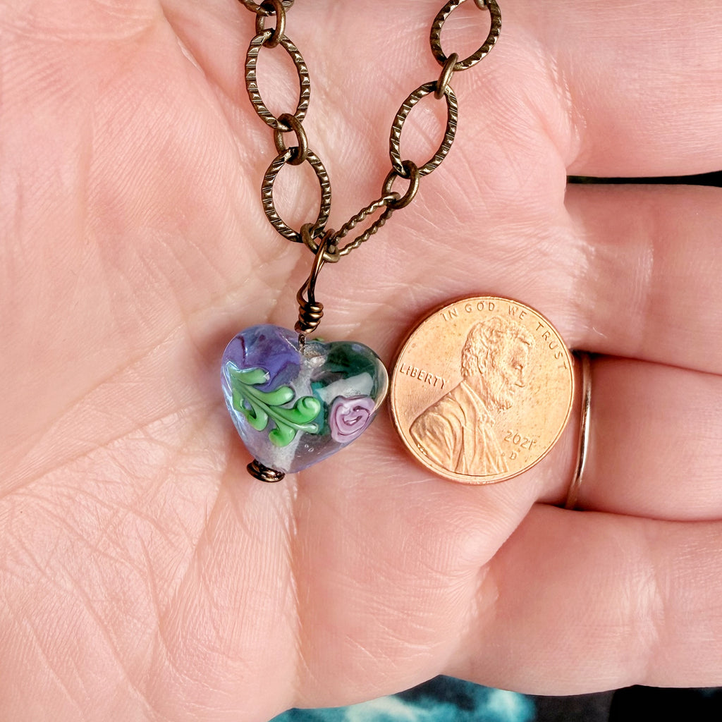 A close-up of a hand holding a heart-shaped glass pendant necklace with a bronze chain next to a US penny for scale. The pendant is made of colorful glass with purple, green, and white floral patterns.