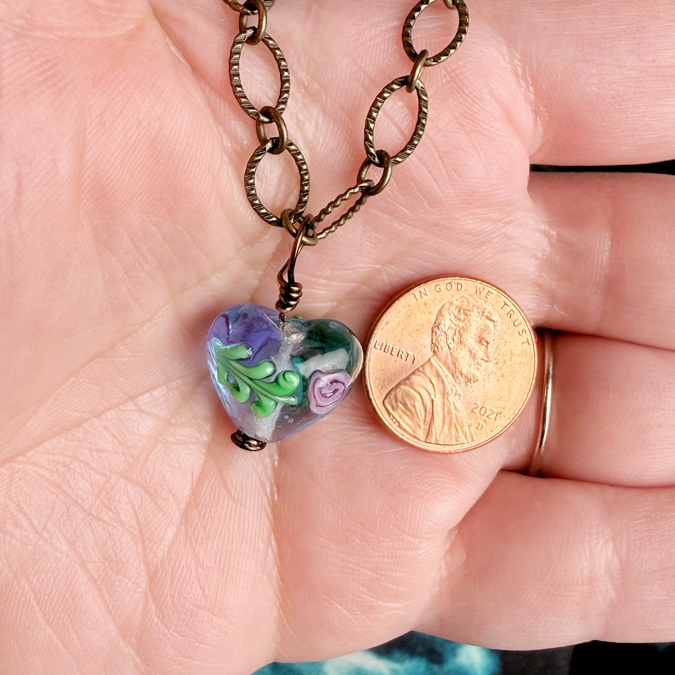 A close-up of a hand holding a heart-shaped glass pendant necklace with a bronze chain next to a US penny for scale. The pendant is made of colorful glass with purple, green, and white floral patterns.