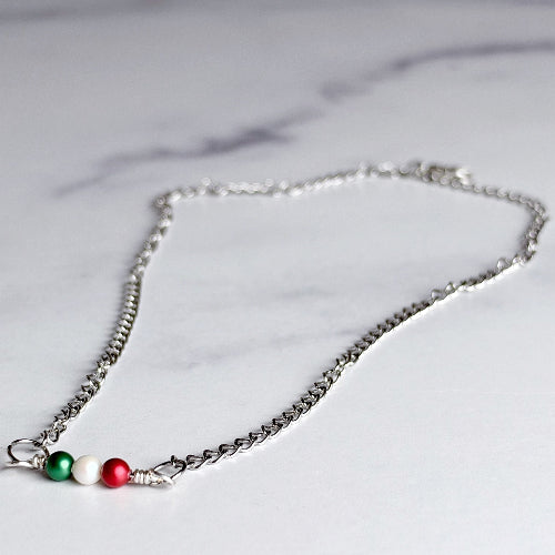 A silver chain necklace with three small decorative beads in green, white, and red, arranged like the Italian flag, resting on a light gray marble surface.