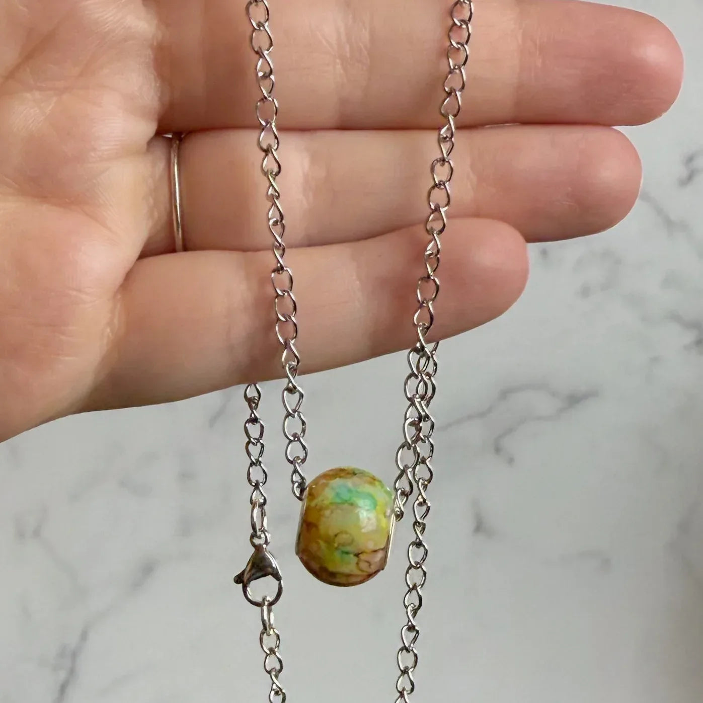 Hand holding a silver chain necklace with a green bead against a marble background