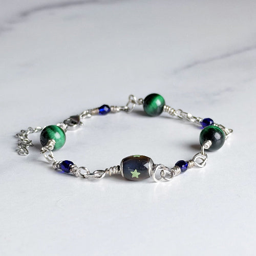 A beaded charm bracelet featuring green malachite beads, a central dark brown bead with a green star motif, and small blue beads, all linked with silver-toned metal chains on a light marble background.