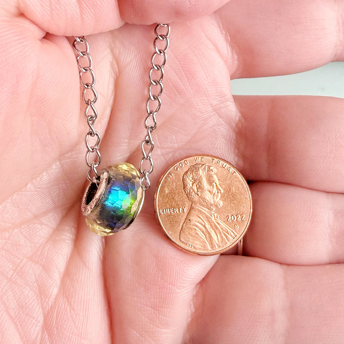 A close-up of a hand holding a small silver chain necklace with an iridescent glass bead pendant next to a U.S penny for scale.