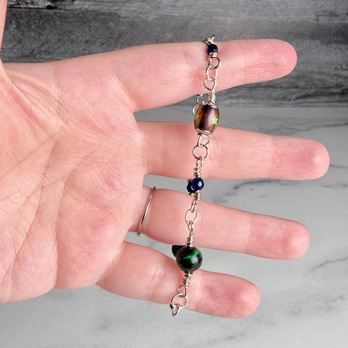 A handmade silver chain bracelet with a large brown and gold iridescent bead, a dark blue bead, and a green bead, held in a person's hand against a gray marble background.