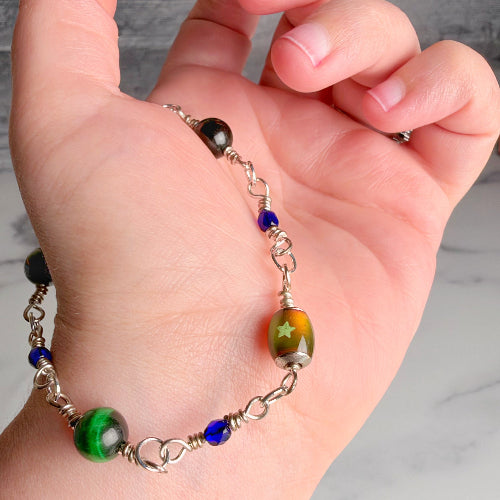 A close-up of a hand holding a handmade beaded bracelet. The bracelet features a silver-toned chain with various decorative beads, including a prominent green and yellow starburst lamp bead, a green tiger-eye style bead, a dark black bead, and small blue accent beads.