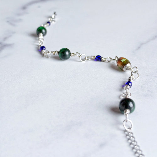 A beaded chain necklace with silver-toned metal links, featuring dark green marbled beads, small blue faceted beads, and a larger iridescent green and orange bead, displayed on a light marble surface.