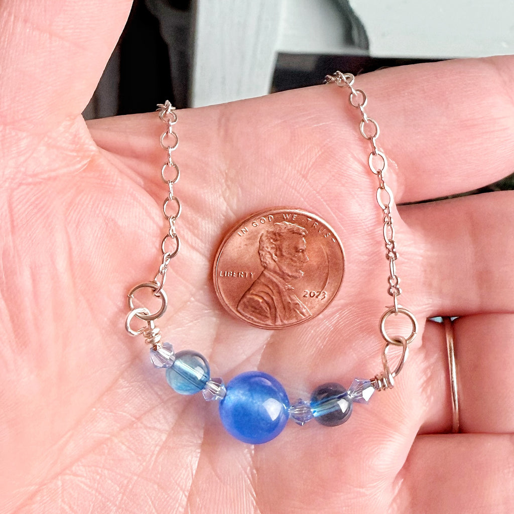 A silver chain bracelet with blue beads, held in a person's hand next to a 2023 US penny for scale.