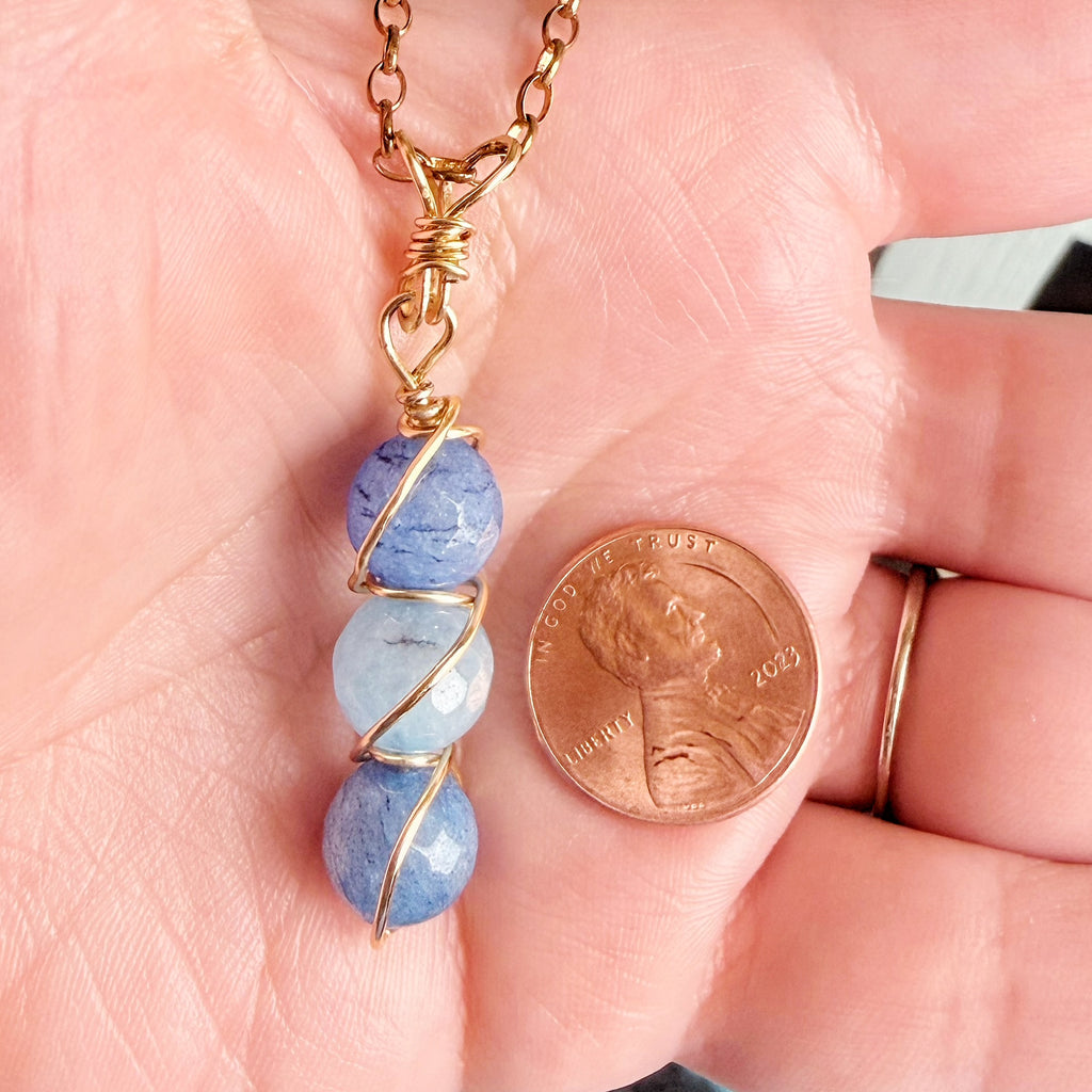 A close-up of a handmade necklace with three blue stone beads wrapped in gold wire, held in a person's hand next to a US penny for scale.