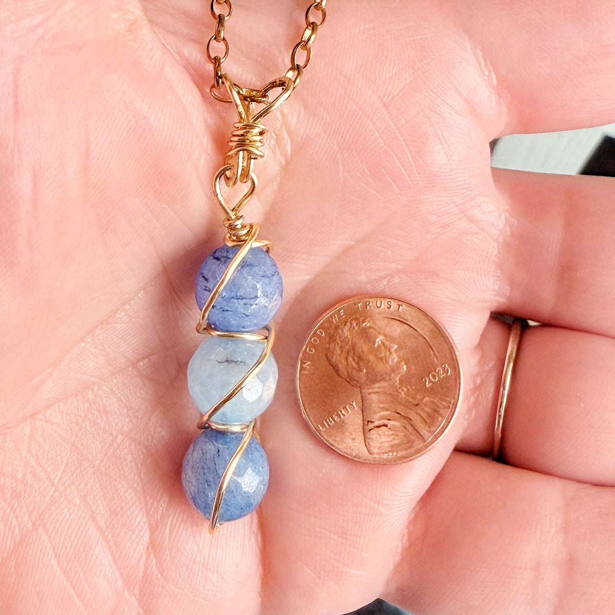 A close-up of a handmade necklace with three blue stone beads wrapped in gold wire, held in a person's hand next to a US penny for scale.