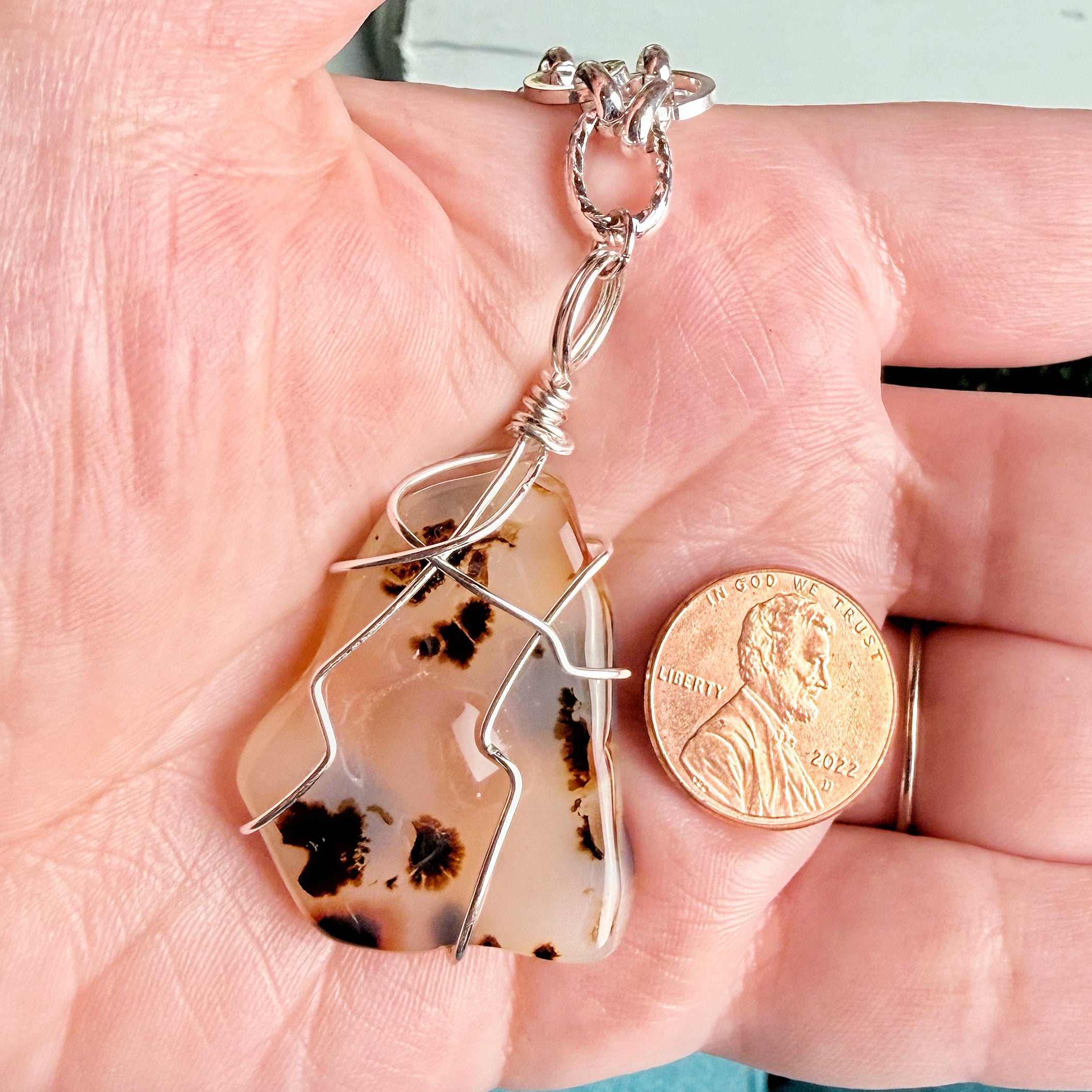 A close-up of a hand holding a silver-wrapped agate pendant next to a US penny for scale, showing the rough stone's natural patterns.