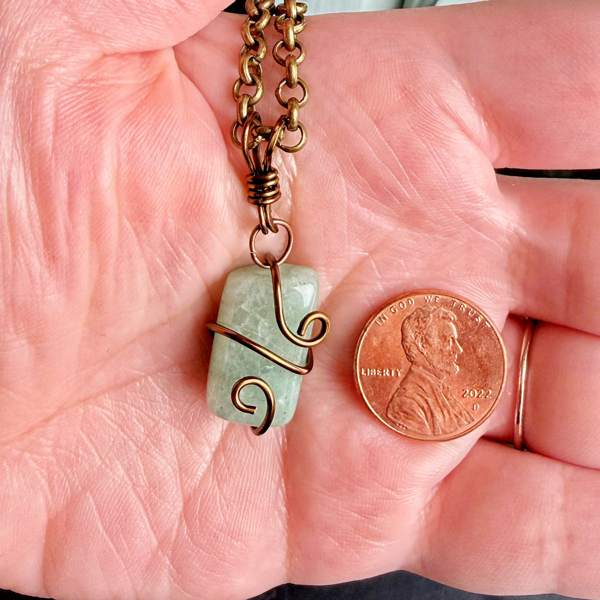 A handmade necklace with a light green rectangular stone wrapped in copper wire and a bronze chain, held in a person's hand next to a US penny for scale.