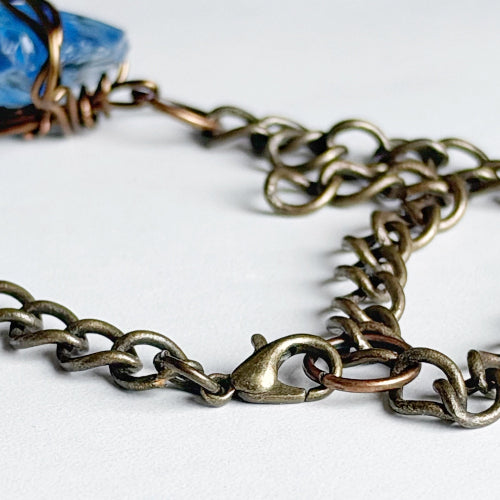 Close-up of an antique bronze chain necklace with a lobster clasp, draped on a white surface with a blue stone and copper wire wrapping visible in the upper left.