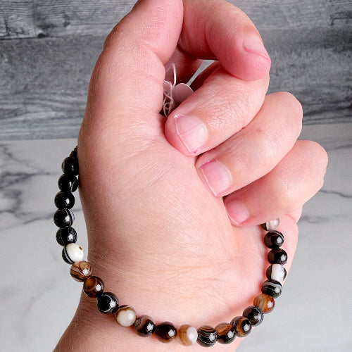 A close-up of a hand wearing a beaded bracelet made of glossy round beads in black, brown, white, and tiger-eye patterns, with a small pink flower charm.
