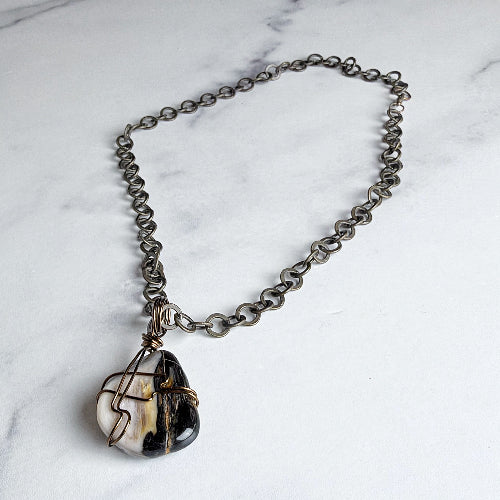 A handmade silver-toned chain necklace with a raw, polished black and white agate stone pendant wrapped in thin copper wire, displayed on a white marble surface.