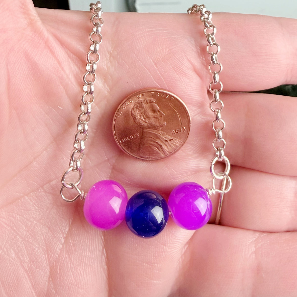 A close-up of a silver chain necklace draped across a person's palm, with three round beads hanging from the chain. The center bead is dark blue, and the two side beads are bright pink.