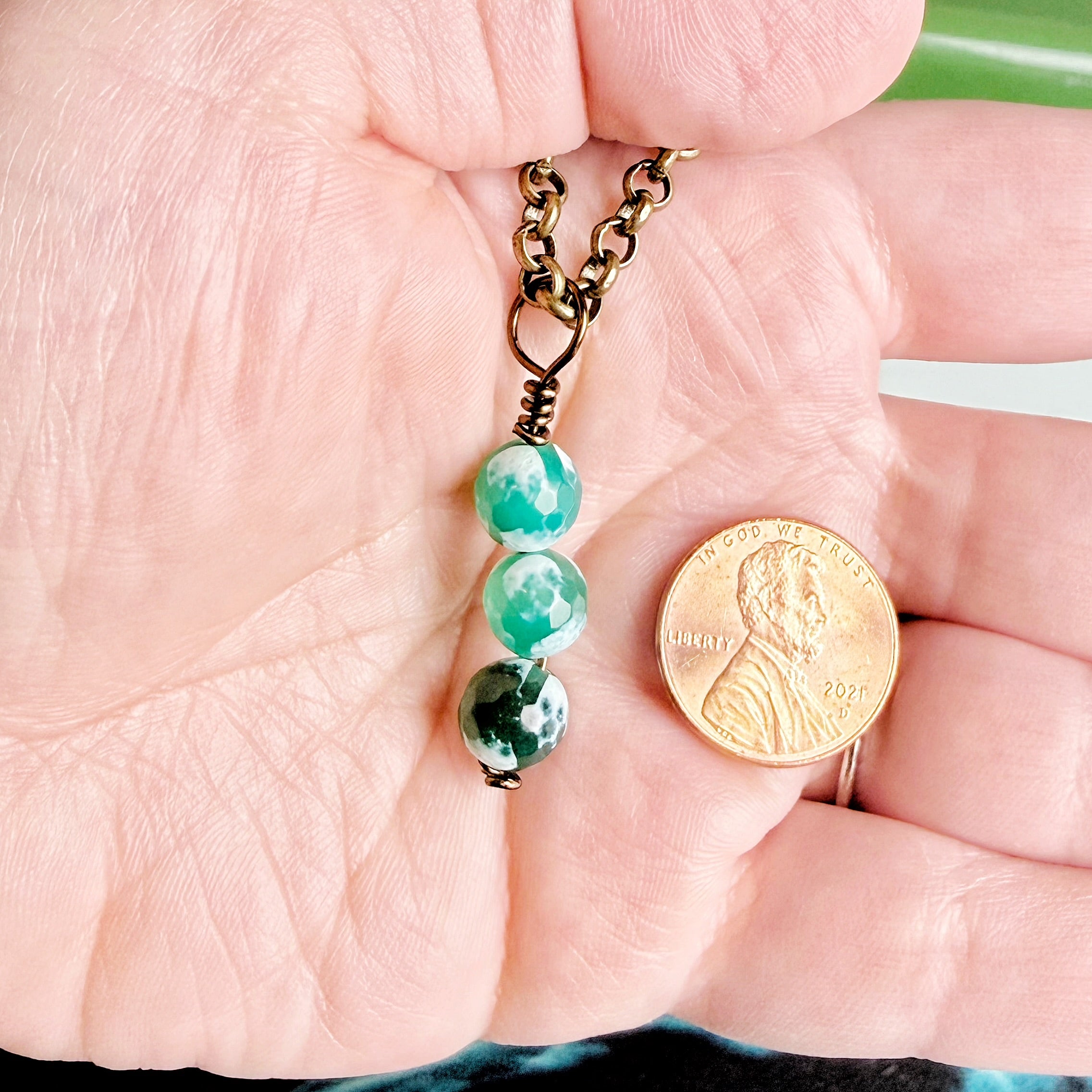 A close-up of a hand holding a small beaded necklace with three green marbled beads and a gold-toned chain, shown next to a U.S penny for scale.