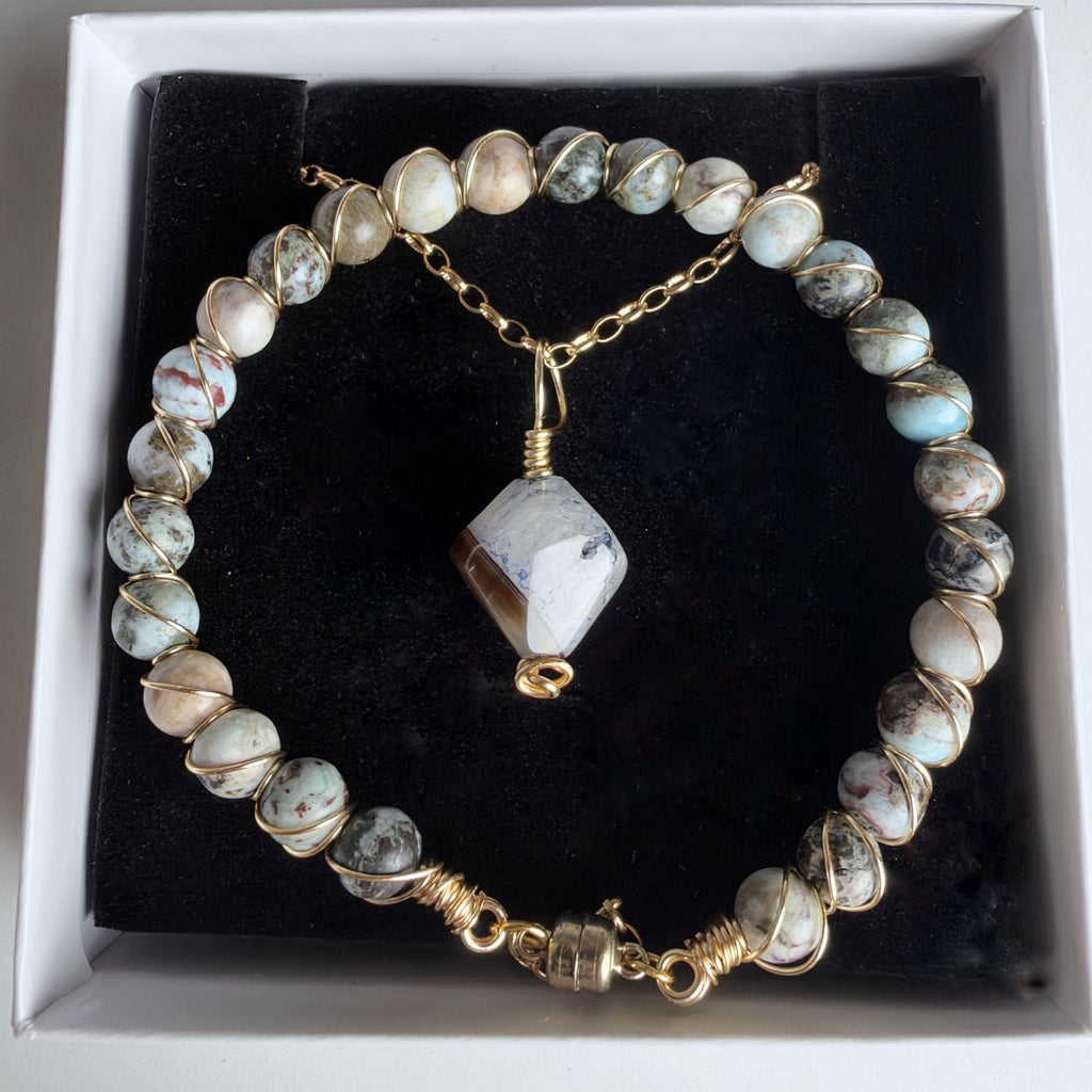 A beaded bracelet with round marbled beads in cream, gray, and light blue, wrapped in gold wire, with a faceted stone pendant and clasp, displayed in a white jewelry box with a black insert.