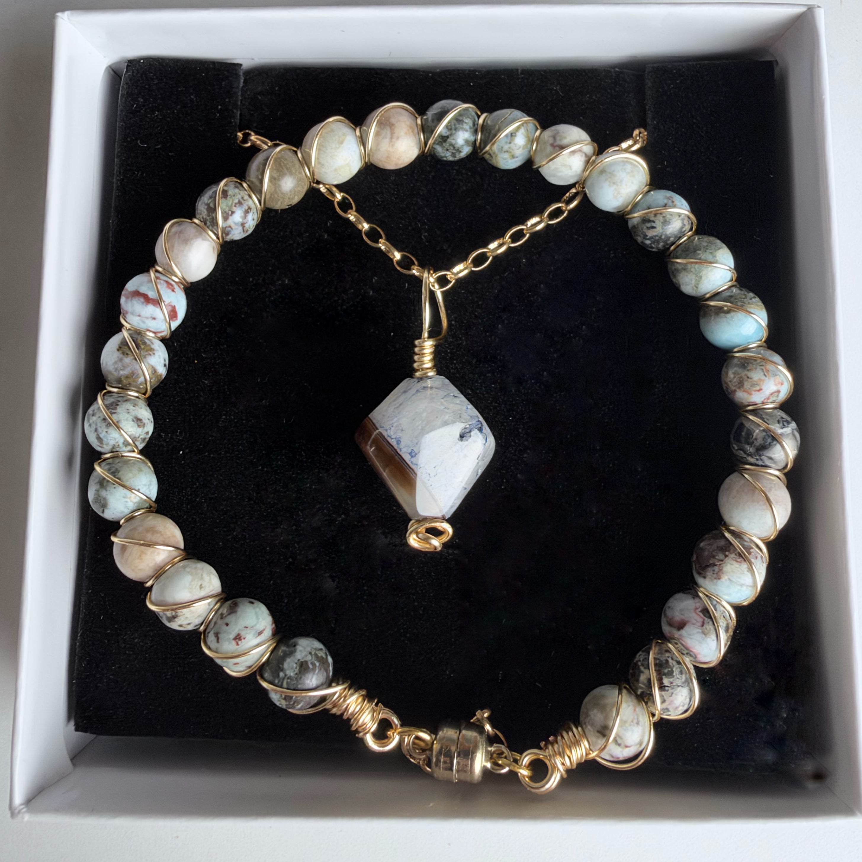 A beaded bracelet with round marbled beads in cream, gray, and light blue, wrapped in gold wire, with a faceted stone pendant and clasp, displayed in a white jewelry box with a black insert.