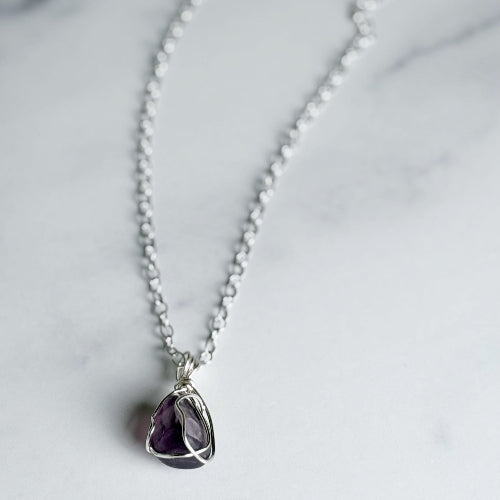 A silver chain necklace with a teardrop-shaped purple amethyst pendant, displayed on a light gray marble surface.