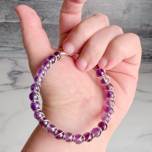 A close-up of a hand holding a beaded bracelet made of purple gemstone beads wrapped in silver-toned metal, against a gray and white marble background.