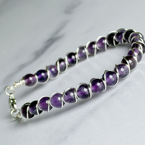 A beaded bracelet featuring polished purple amethyst gemstones in a spiral wrap design with silver-toned metal, resting on a light gray marble surface.