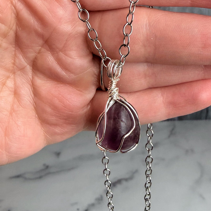 Hand holding a silver necklace with a purple pendant against a marble background