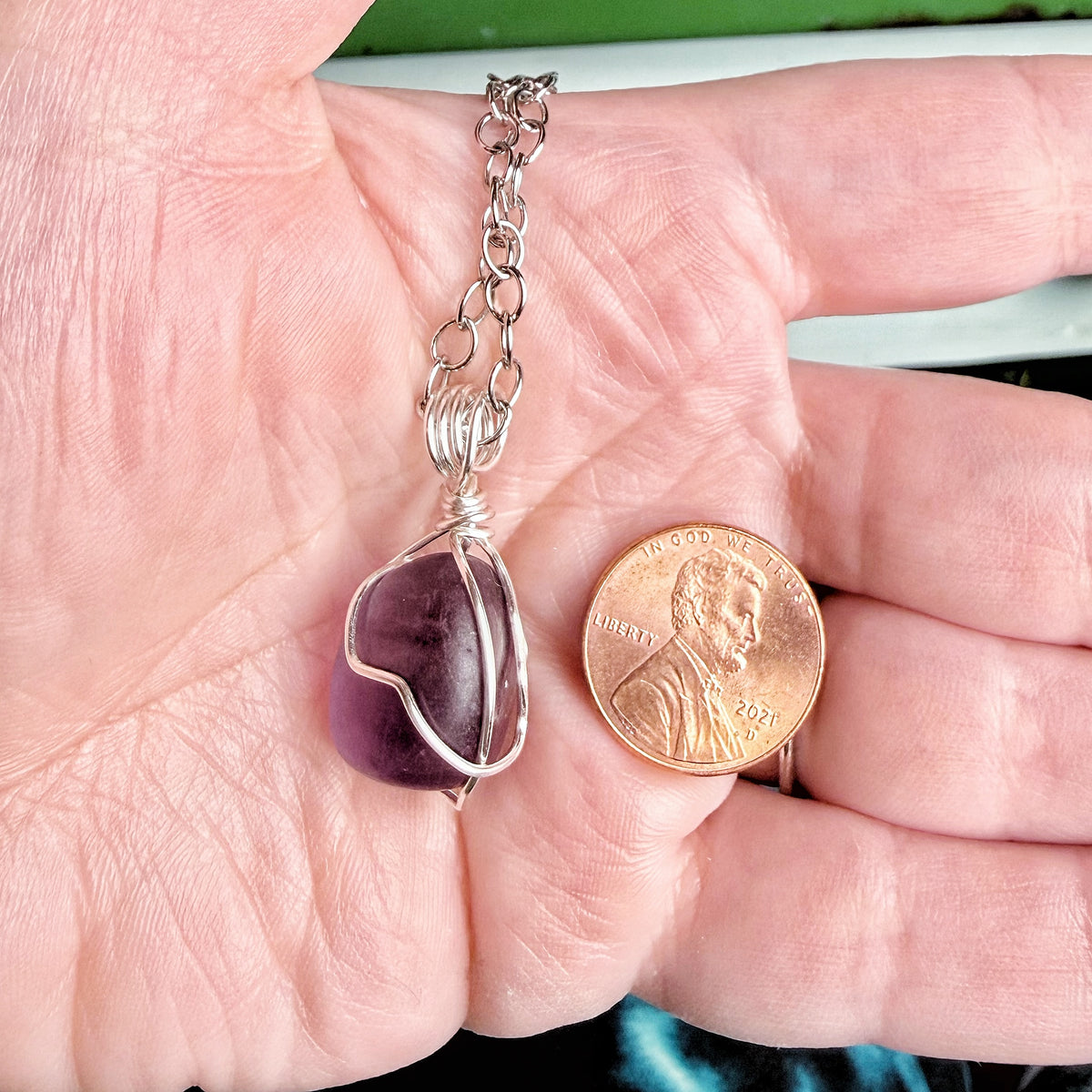 A hand holding a silver chain necklace with a purple stone wrapped in silver wire, shown next to a U.S. penny for scale.