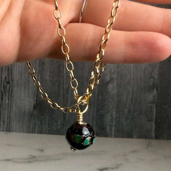 Gold chain necklace with a black bead held by a hand against a marble background