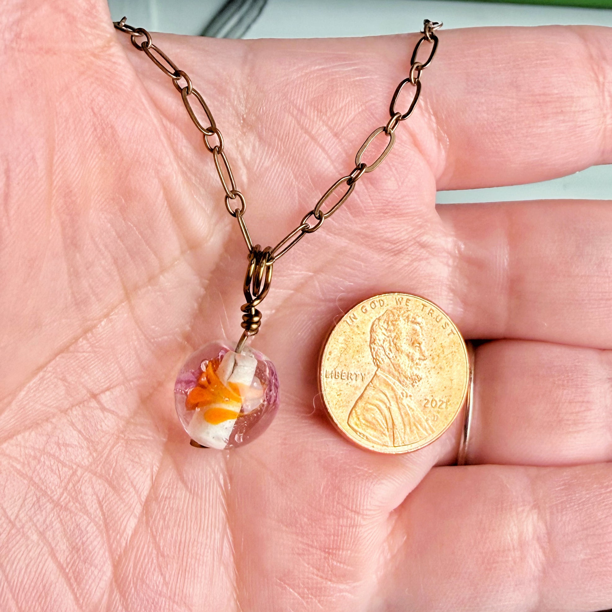 A close-up of a bronze chain necklace with a small clear heart-shaped pendant containing orange and white flower-like inclusions, displayed on an open hand next to a U.S penny for scale.