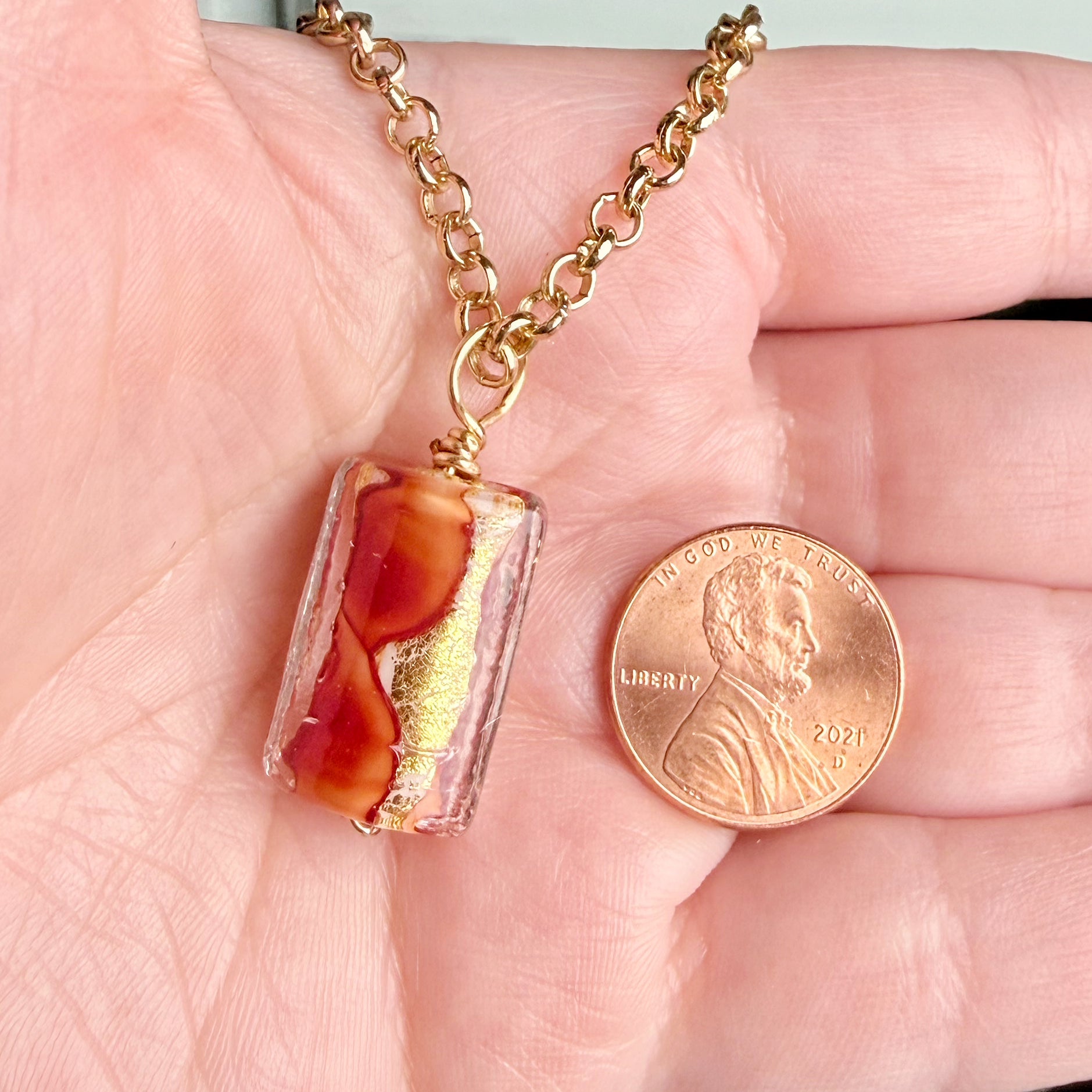 A close-up of a hand holding a gold chain necklace with a rectangular pendant containing amber and orange inclusions, shown next to a U.S penny for scale.