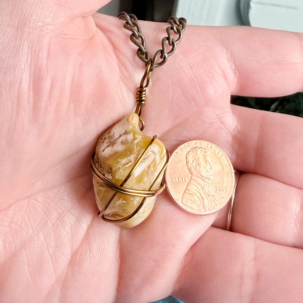A close-up of a hand holding a raw, polished yellow stone pendant wrapped in gold-toned wire, next to a US penny for scale, with a dark metal chain attached.
