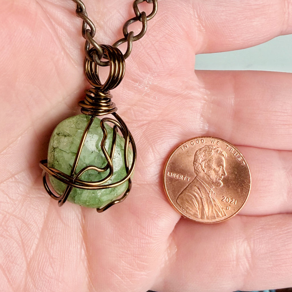 A close-up of a handmade wire-wrapped pendant necklace with a green stone, held in a person's hand next to a US penny for scale.