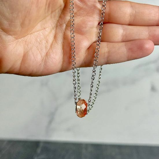 Hand holding a silver chain with a small orange pendant against a neutral background
