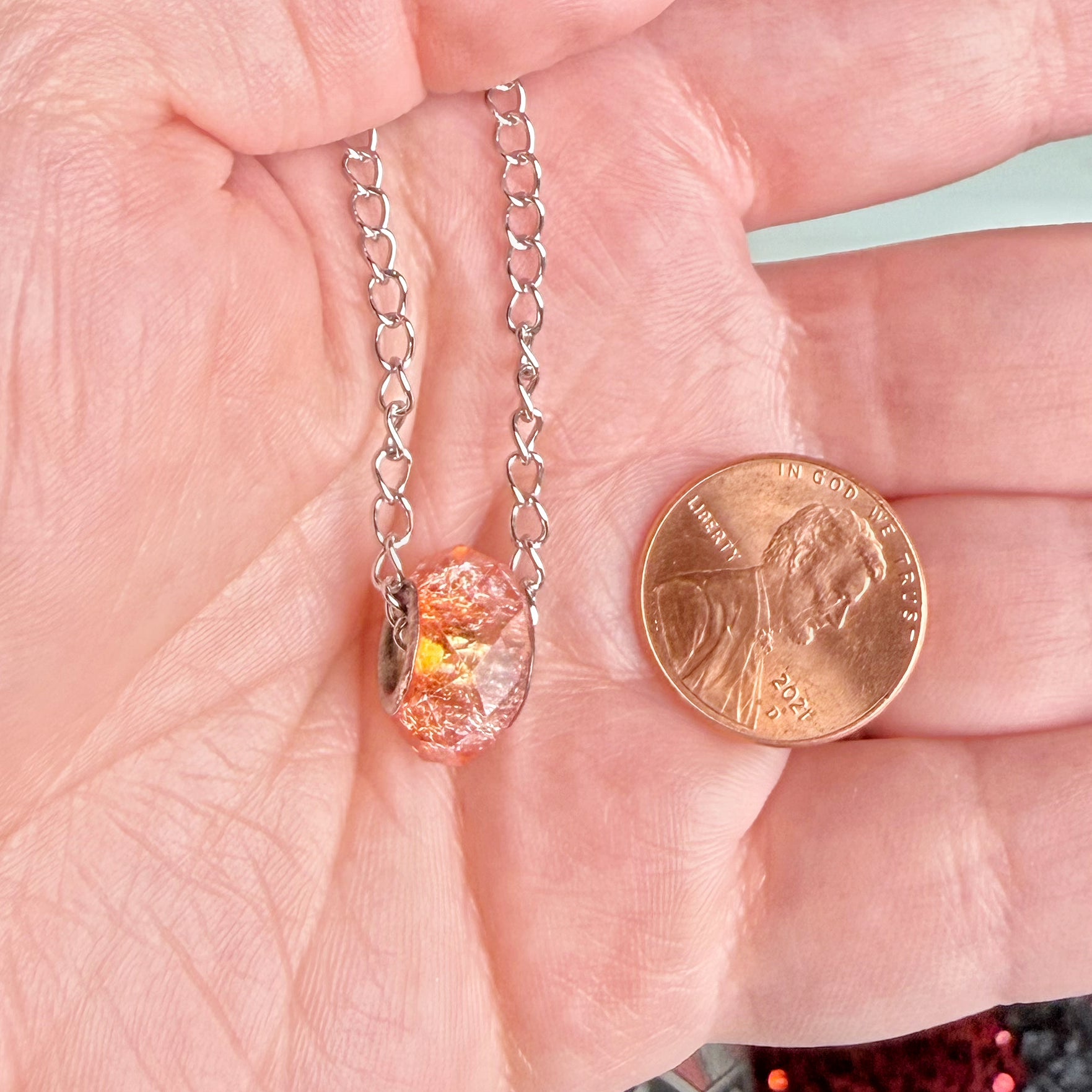 A close-up of a hand holding a silver chain necklace with a small, irregularly shaped orange and yellow glittery pendant, shown next to a US penny for scale.