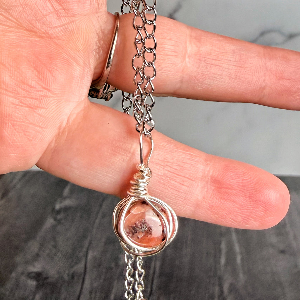 Hand holding a silver necklace with a pendant featuring a peach-colored stone.