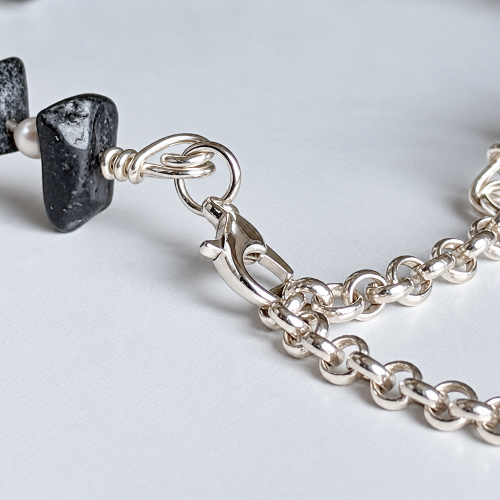 Close-up of a silver chain bracelet with a lobster clasp and a dark polished stone bead, photographed on a light gray background.