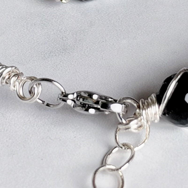 Close-up of a silver wire-wrapped chain bracelet with a lobster clasp and a glossy black bead on a light gray background.