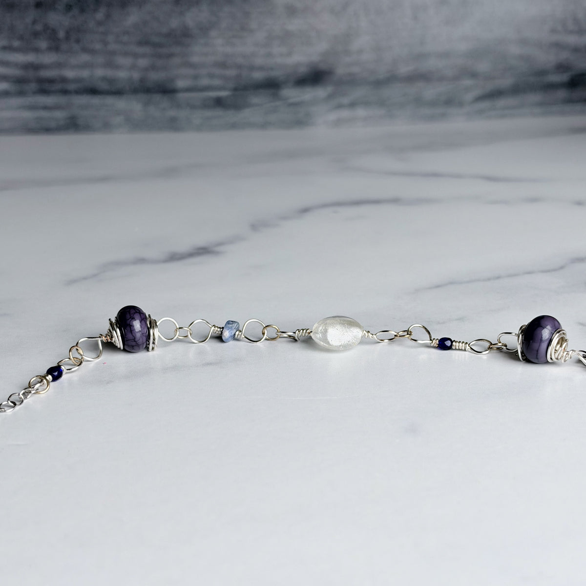 A delicate silver chain bracelet featuring two dark purple glass beads, a central clear glass bead, and small blue beads, displayed on a white marble surface.