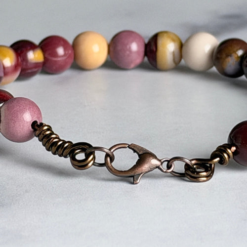 A close-up of a beaded jewelry strand with round beads in pink, cream, yellow, and brown marbled stone, attached to an antique bronze lobster clasp on a light gray background.