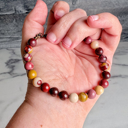 A beaded stretch bracelet made of polished round stones in red, burgundy, cream, yellow, and marbled pink and white tones, shown worn on a person's hand against a gray wood-and-marble background.