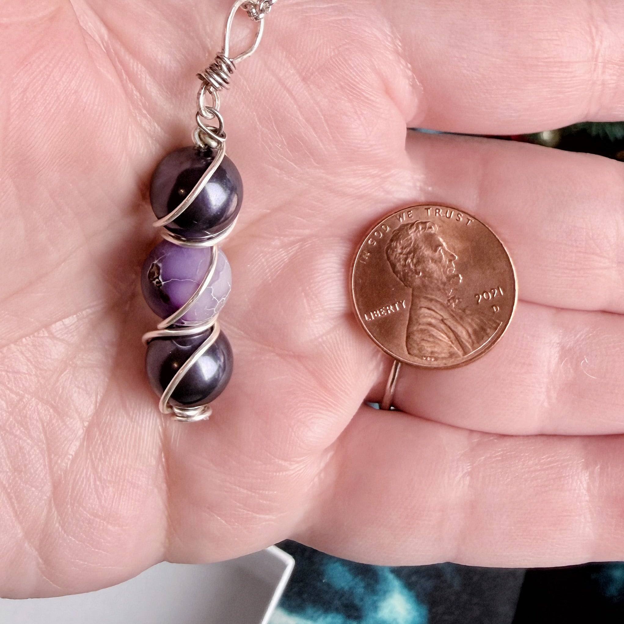 A close-up of a hand holding a silver-wrapped beaded charm or pendant next to a U.S penny for scale