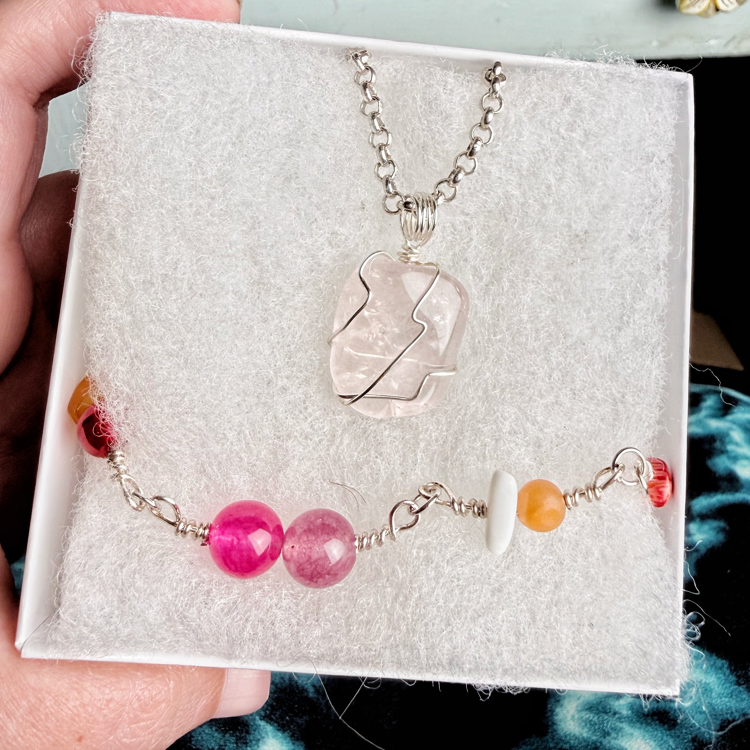 A gift set in a white box with a silver chain necklace featuring a clear crystal pendant and a matching beaded bracelet with pink, orange, and white beads, held by a hand.