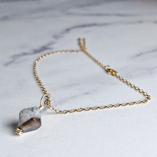 A delicate gold chain necklace featuring a small, irregular faceted stone pendant with a white and gray marbled top half and a smooth brown bottom half, displayed on a light gray marble surface.