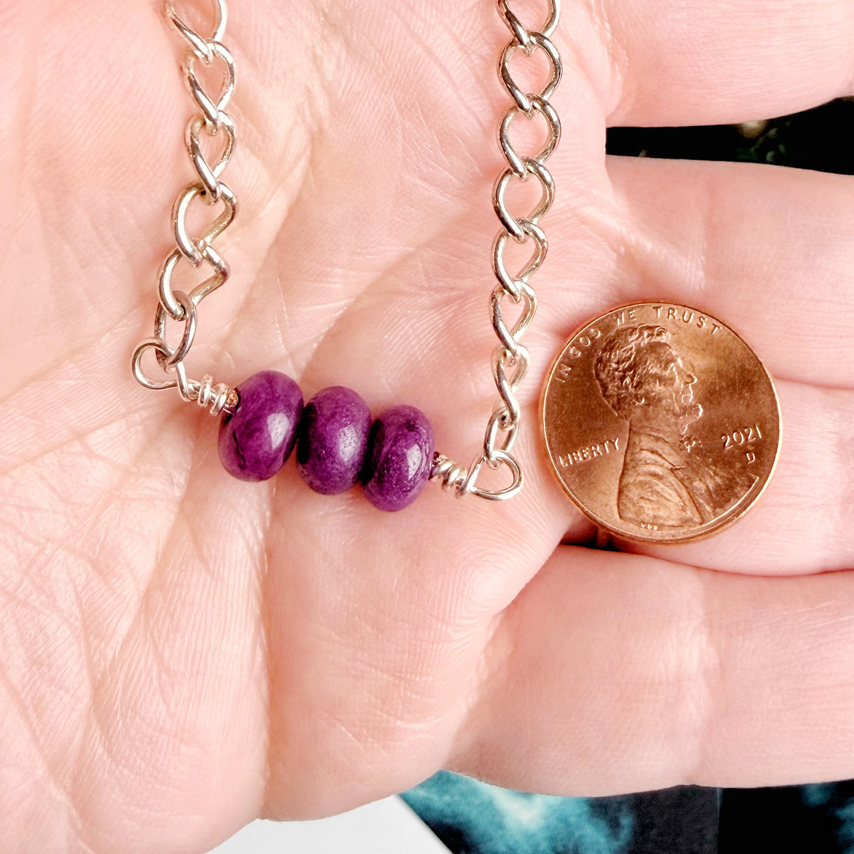 A close-up of a silver chain necklace with three purple beads, held in a person's hand next to a U.S. penny for scale.